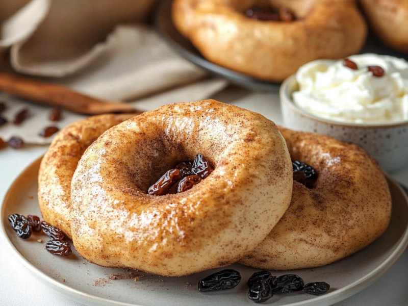 Ein Teller mit 3 Rosinen-Zimt-Bagel, daneben ein Messer und eine Schale Frischkäse, dahinter weitere Bagel.
