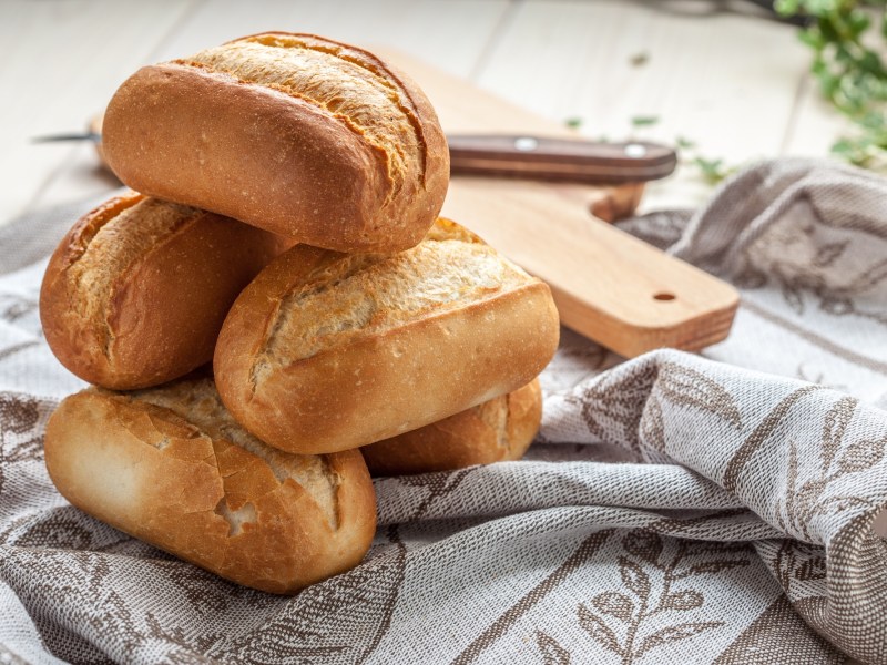 Ein paar Sonntagsbrötchen auf einem Geschirrtuch, dahinter ein Holzbrett.