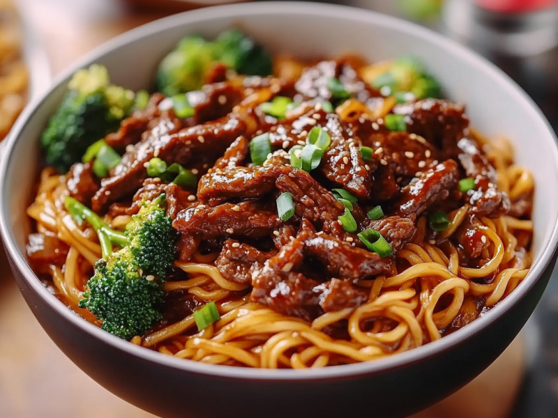 Eine Schale mit Sticky Beef Noodles mit Brokkoli und Frühlingszwiebeln. Daneben liegen Stäbchen und einige Zutaten.