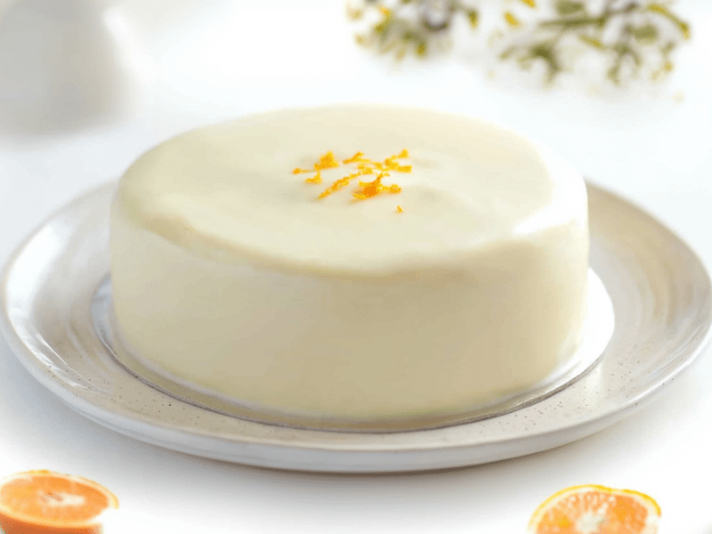 Ein großer Teller auf dem eine ganze weiße Sachertorte mit Orangenmarmelade liegt. Die weiße Sachertorte ist mit ein kleines bisschen Orangenabrieb dekoriert. Um den Teller herum liegen eine ganze Orange und zwei Orangenhälften. Außerdem kann man verschwommen Zweige mit Blättern und Blüten erkennen. Der Teller mit der Torte steht auf einem weißen Untergrund.