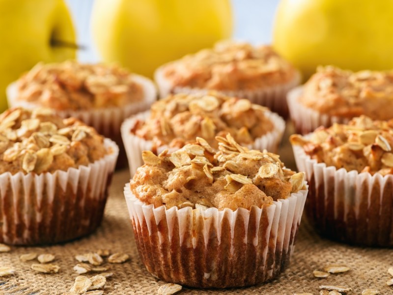 Apfel-Haferflocken-Muffins auf Holz, dahinter gelbe Äpfel.