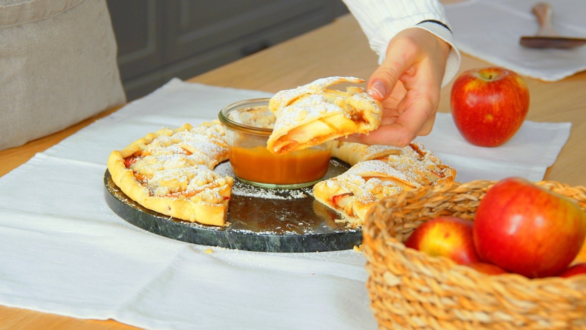 Eine ringförmige Apfeltarte mit Lotus-Creme, mit einer Schüssel Karamell-Dip in der Mitte. Der Ring ist angeschnitten und es ist eine Hand zu sehen, die sich ein Stück der Apfeltarte mit Lotus-Creme nimmt. Die Tarte ist mit Puderzucker bestäubt. Das runde Blech mit der Tarte steht auf einer Holzarbeitsfläche, die mit einem weißen Tuch bedeckt ist. Außerdem ist ein Korb mit Äpfeln zu sehen.