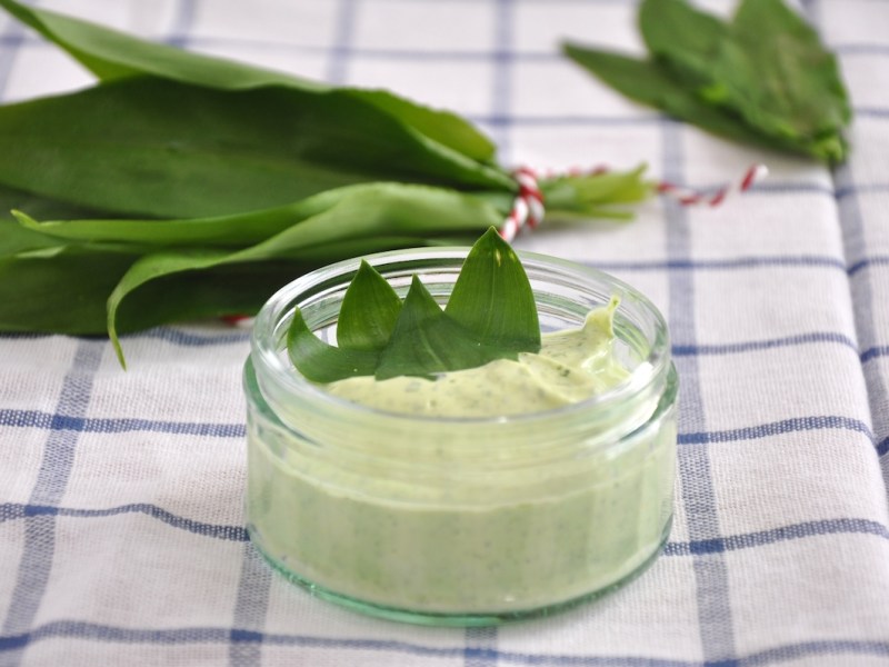Eine kleine Glasschale Bärlauch-Curry-Aufstrich garniert mit Bärlauchblättern, im Hintergrund mehr Bärlauch.