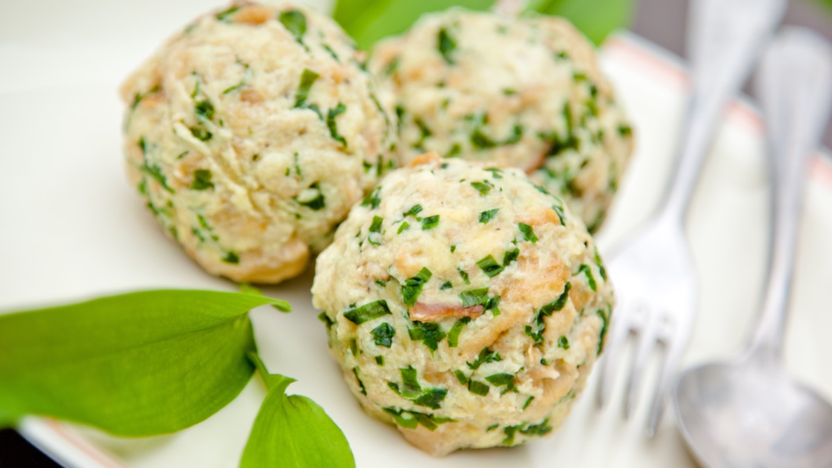 Drei Bärlauchknödel auf einem Teller. Daneben liegen Besteck und frischer Bärlauch.