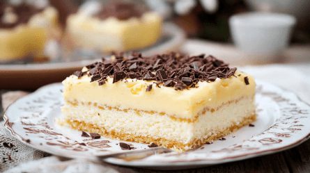 Ein Teller mit einem rechteckigen Stück Eierlikörkuchen vom Blech. Der Kuchen hat eine Eierlikörpuddingschicht und ist mit Schokostückchen getoppt. Im Hintergrund kann man weitere Stückchen Eierlikörkuchen vom Blech erkennen.
