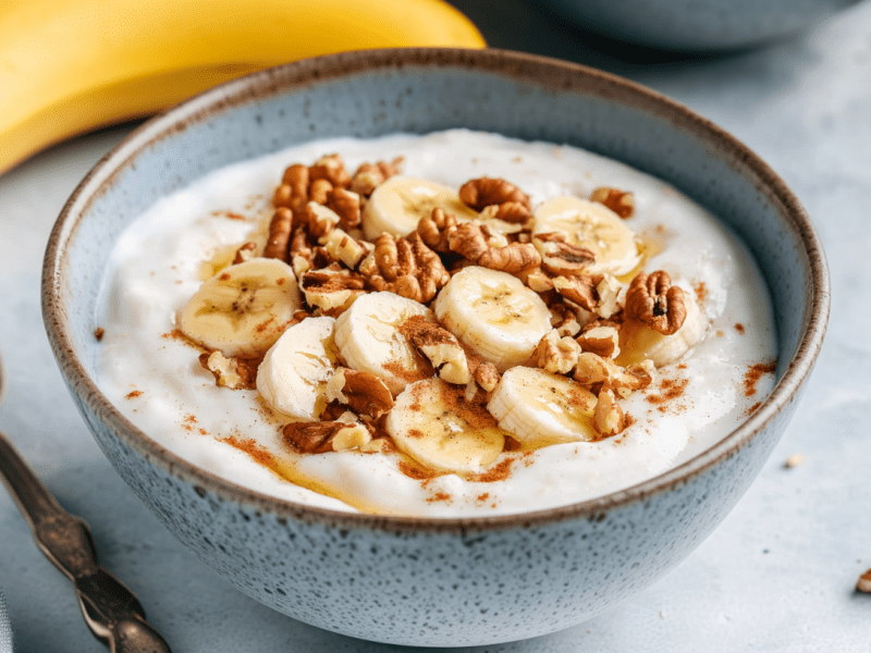 Eine Schale Frühstücks-Skyr mit Nüssen und Honig. Zusätzlich sind ein paar Bananenscheiben in der Schale verteilt. Im Hintergrund liegen zwei Bananen. Links neben der Schale ein Löffel. Rechts ein paar gehackte Nüsse.
