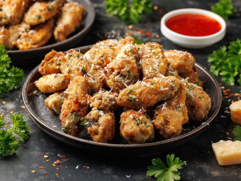 Ein Teller Knoblauch-Parmesan-Hühnchen aus dem Airfryer, daneben eine Schale mit roter Salsa und Parmesanstücke.