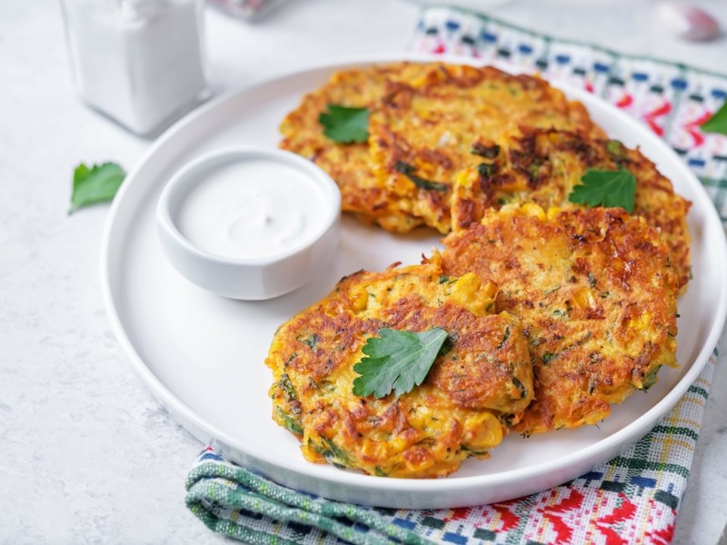 Ein paar Maisfrikadellen auf einem Teller, auf dem auch ein Schälchen mit Schmand steht. Der Teller steht auf einem Geschirrtuch.