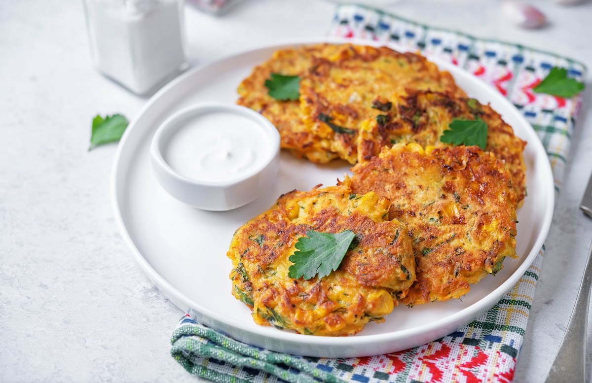 Ein paar Maisfrikadellen auf einem Teller, auf dem auch ein Schälchen mit Schmand steht. Der Teller steht auf einem Geschirrtuch.
