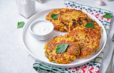 Ein paar Maisfrikadellen auf einem Teller, auf dem auch ein Schälchen mit Schmand steht. Der Teller steht auf einem Geschirrtuch.