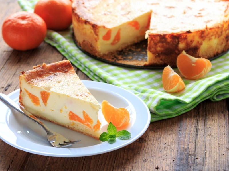 Ein Stück Mandarinen-Quark-Kuchen auf einem Teller, daneben der Kuchen und ein paar Mandarinen.