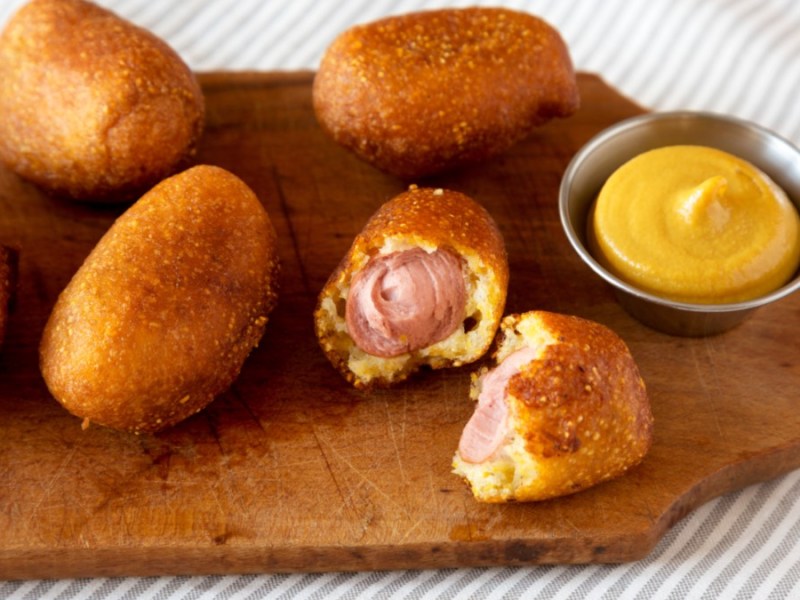 Mehrere Mini Corn Dogs aus dem Airfryer auf einem Holzbrett. Daneben steht ein Schälchen mit gelbem Dip.