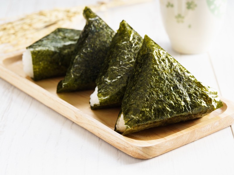 Vier Onigiri liegen auf einem kleinen Holzbrett. Im Hintergrund sieht man eine weiße Tasse.