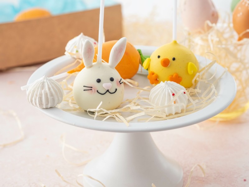 Osterhasen-Cake-Pop und ein Küken-Cake-Pop auf einer Tortenplatte mit weiterer Deko.