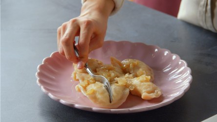 Ein rosa Teller mit einer Portion selbst gemachter Pierogi. Man kann eine Hand erkennen, die eine Gabel hält und eine Pirogge zerteilt.