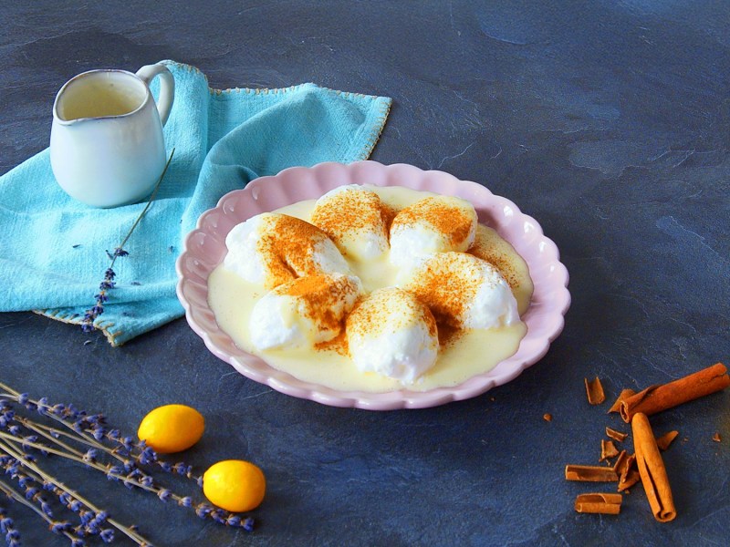 Ein rosa Teller mit Farófias. Dahinter liegt ein blaues Tuch auf dem eine kleine Kanne steht. Im Vordergrund liegen noch einige kleine Zwiebeln, Zimtstangen und Blumen.