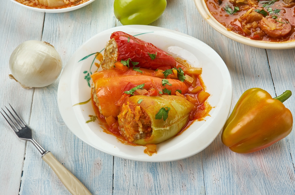 3 ungarische gefüllte Paprika mit Tomatensoße auf einem Teller, daneben eine Gabel, eine Paprika und eine Zwiebel sowie ein Topf mit Tomatensoße.