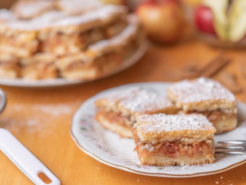 Ein paar Stücke ungarischer Apfelkuchen auf einem Kuchenteller, im Hintergrund weitere Stücke und Äpfel.