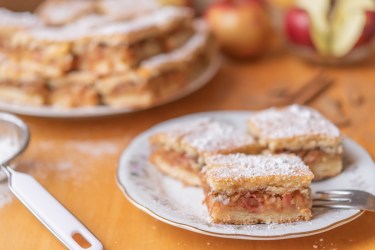 Ein paar Stücke ungarischer Apfelkuchen auf einem Kuchenteller, im Hintergrund weitere Stücke und Äpfel.