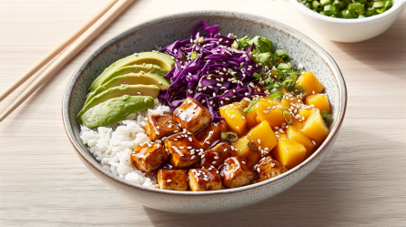 Eine Veggie Teriyaki-Bowl steht auf einem Holztisch. Sie besteht aus Reis, Tofuwürfeln, Mango, Frühlingszwiebeln, Rotkohl und Avocado. Links von ihr liegen Essstäbchen, rechts im Hintergrund sieht man eine Schale Frühlingszwiebeln.