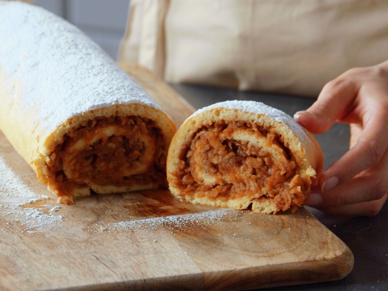 Ein Holzbrett, auf dem eine süße Apfelrolle liegt, die mit Puderzucker bestäubt ist. Man kann eine Hand erkennen, die ein abgeschnittenes Stück präsentiert.