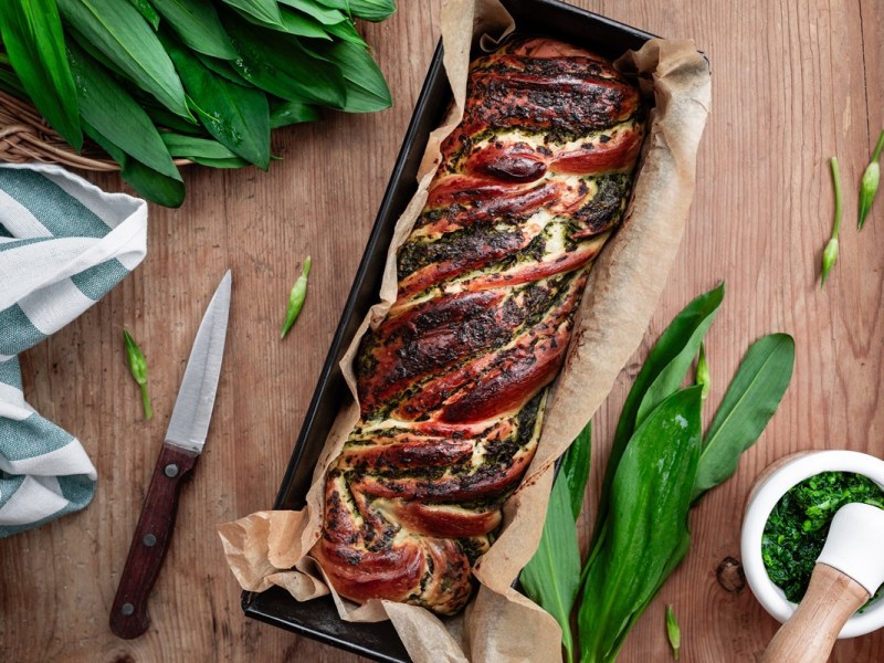 Bärlauch-Hefezopf in einer Kastenform. Daneben liegt Bärlauch sowie ein Messer.