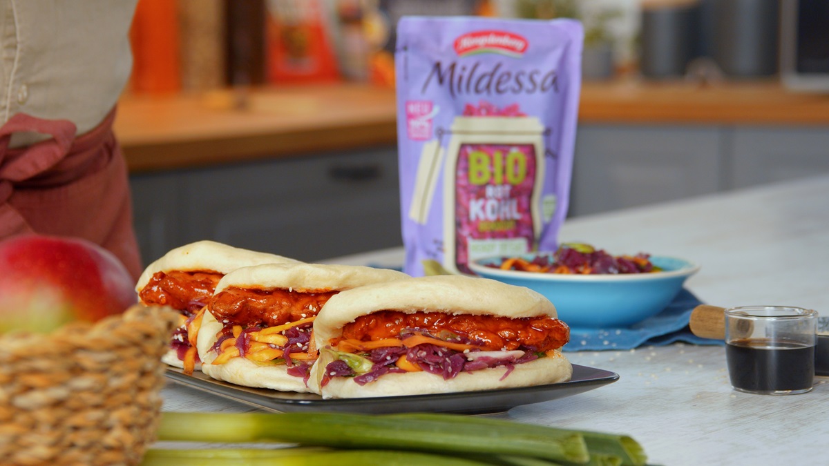 Bao Buns mit Rotkohl-Mangosalat und Hähnchen auf einem Teller. Im Hintergrund ist eine Tüte MIldessa Bio Rotkohl gekocht zu sehen.