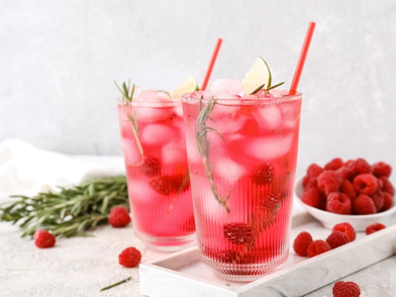 2 Gläser Berry-Lime-Cocktail mit Himbeeren, Eiswürfeln, Limettenspalten und Rosmarin, in denen ein Strohhalm steckt. Daneben steht eine Schale mit Himbeeren.
