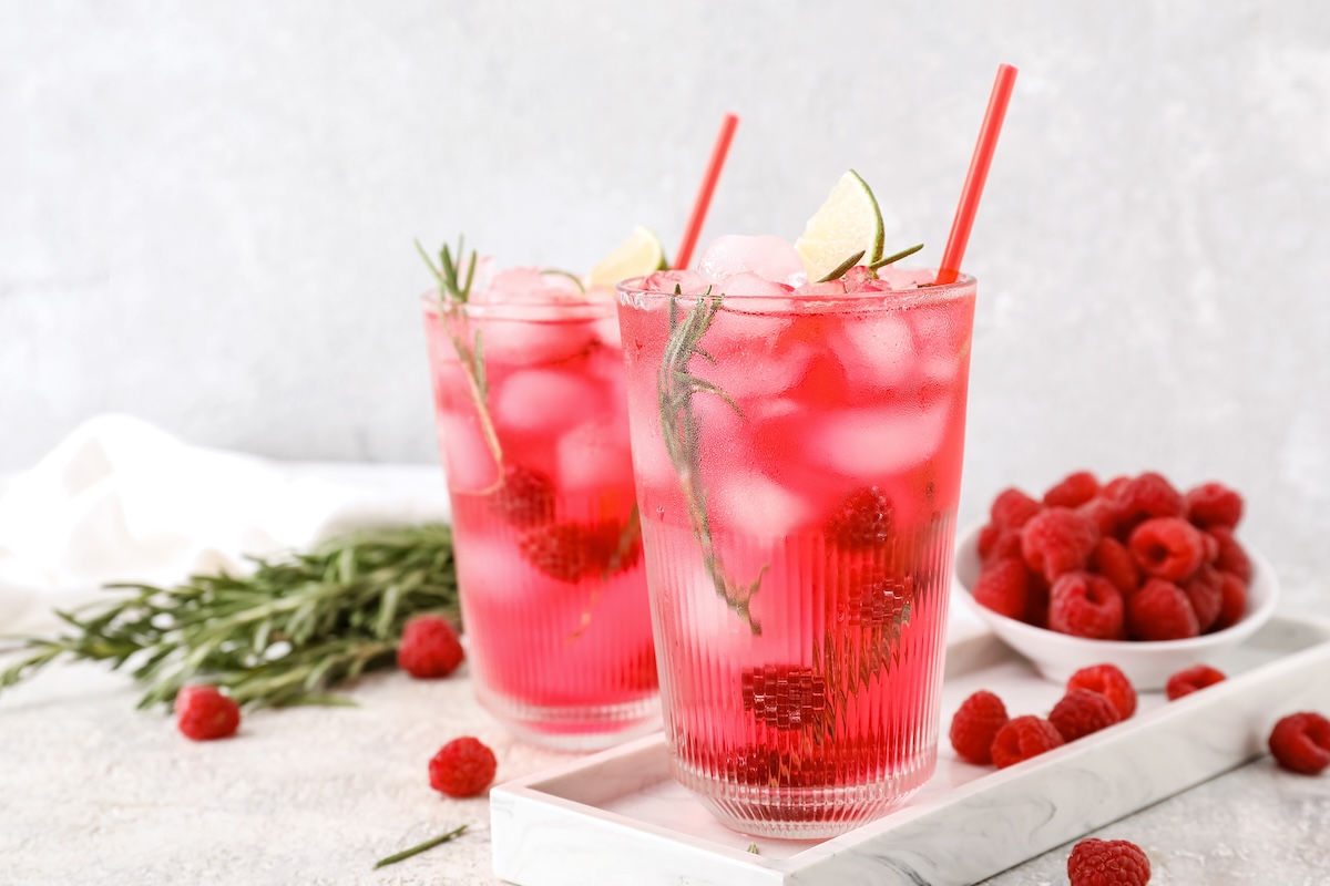 2 Gläser Berry-Lime-Cocktail mit Himbeeren, Eiswürfeln, Limettenspalten und Rosmarin, in denen ein Strohhalm steckt. Daneben steht eine Schale mit Himbeeren.