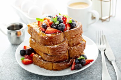 Ein Stapel French Toasts mit Eierlikör, garniert mit frischen Beeren, auf einem Teller. Daneben Besteckt, eine Kanne Sirup und eine Tasse.