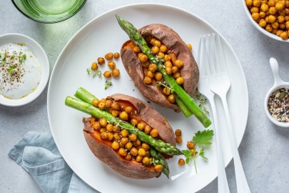 1 halbierte gebackene Süßkartoffel mit Spargel und Kichererbsen auf einem Teller, daneben weißes Besteck und Schüsseln mit Zutaten. Draufsicht.