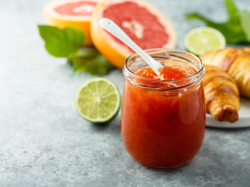Glas mit Grapefruit-Limetten-Marmelade, Grapefruit, Limetten und Croissant daneben.
