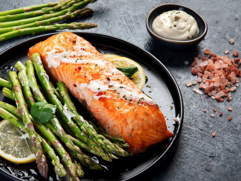 Knoblauch-Zitronen-Spargel aus dem Ofen mit einem Stück Lachsfilet auf einem Teller. Daneben frischer Spargel und eine Schale weißer Dip.