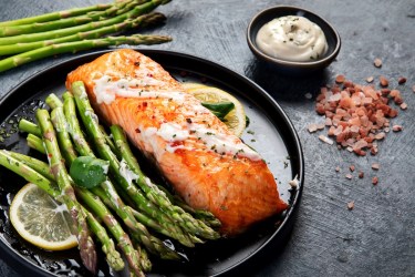 Knoblauch-Zitronen-Spargel aus dem Ofen mit einem Stück Lachsfilet auf einem Teller. Daneben frischer Spargel und eine Schale weißer Dip.