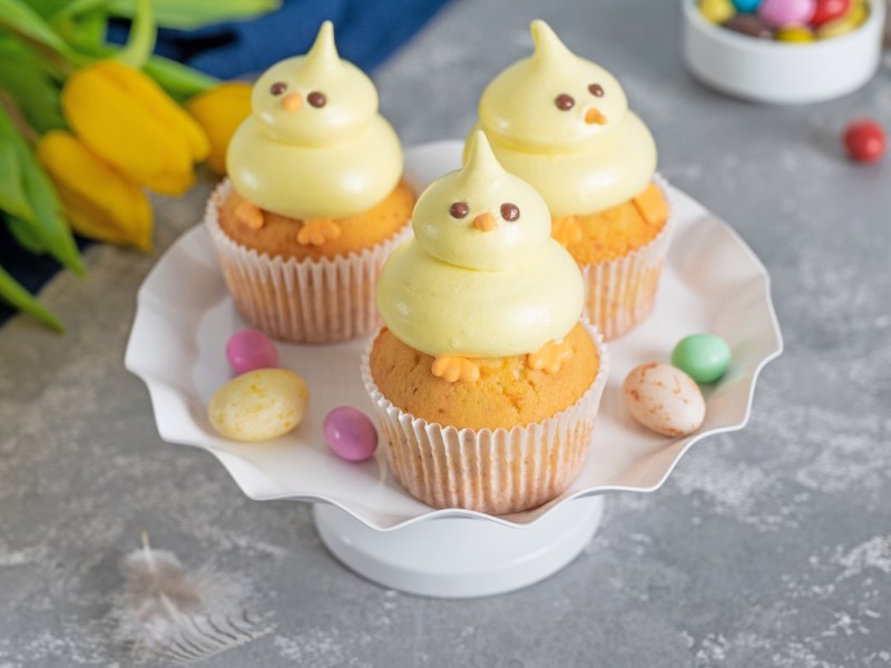3 Küken-Cupcakes auf einem Teller, drumherum Osterdeko und Tulpen.
