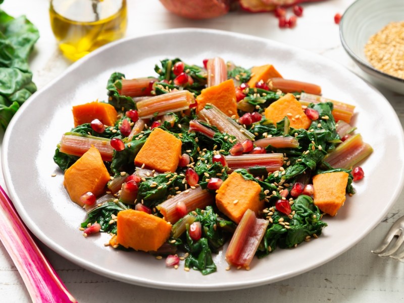Ein Teller lauwarmer Süßkartoffel-Mangoldsalat mit Granatapfelkernen und Sesam, drumherum einige der Zutaten.