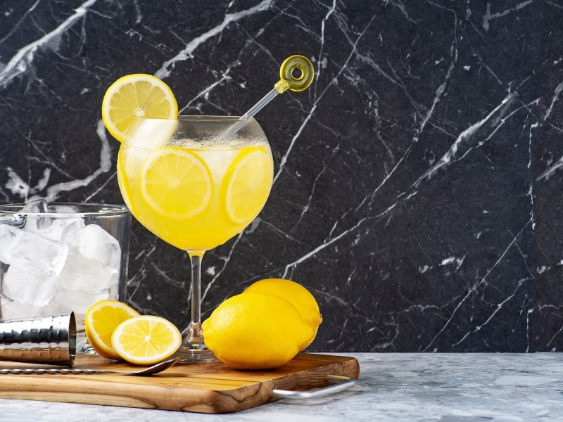 Ein Glas Limoncello Gin Fizz auf einem Brett, daneben Zitronen und ein Glas mit Eiswürfeln.