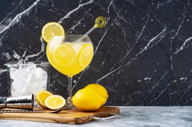 Ein Glas Limoncello Gin Fizz auf einem Brett, daneben Zitronen und ein Glas mit Eiswürfeln.