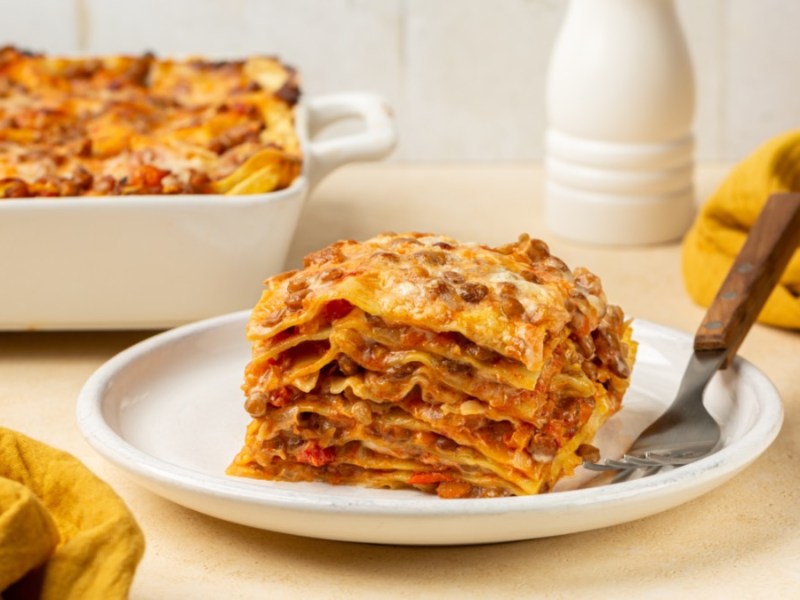 Ein Stück Linsen-Lasagne auf einem Teller. Im Hintergrund ist der Rest in einer Auflaufform zu sehen.
