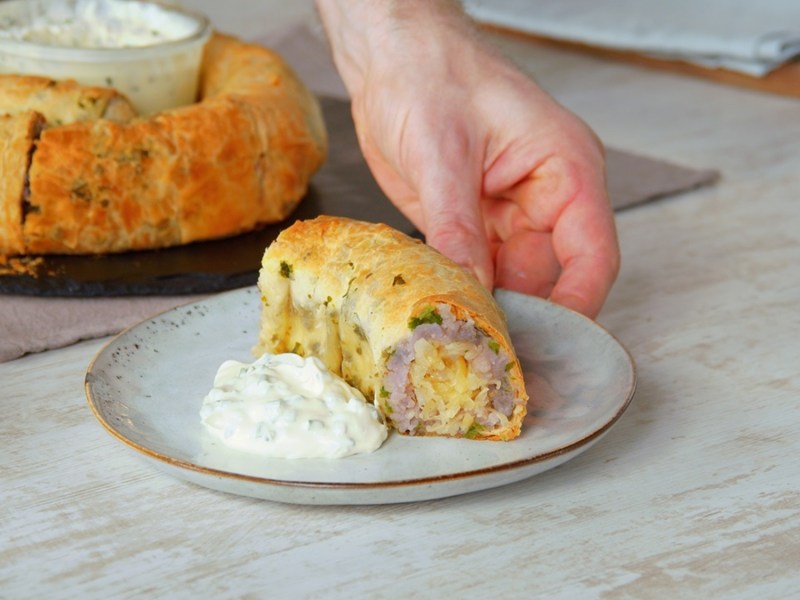 Ein Stück Weisskraut-Kartoffel-Strudel mit einer Portion Sour Cream auf einem Teller. Im Hintergrund kann man den restlichen, schneckenförmigen Strudel erkennen. In der Mitte des Strudels ist eine Schüssel mit Sour Cream platziert.