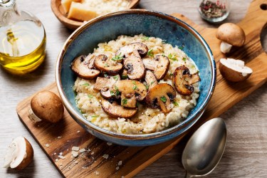 Eine Schüssel Pilzrisotto auf einem Brett, daneben Champignons und ein Löffel.