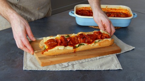 Ein Schaschlik-Sandwich auf einem Holzbrett. Unter dem Brett liegen Tischuntersetzer aus Stoff. Neben dem Schaschlik-Sandwich steht eine hellblaue Auflaufform mit Schaschlik-Soße und weiterem Schaschlik. Man kann zwei Hände erkennen, die nach dem Sandwich greifen.