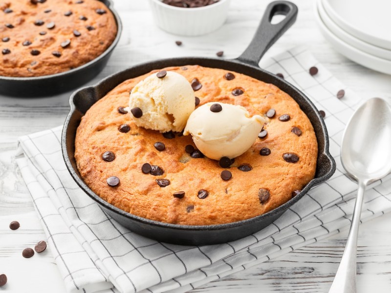 EIn Skillet Cookie, also Pfannenkeks, mit Schokotropfen in einer Pfanne, garniert mit zwei Eiskugeln. Daneben ein Esslöffel und eine weitere Pfanne mit einem Keks.