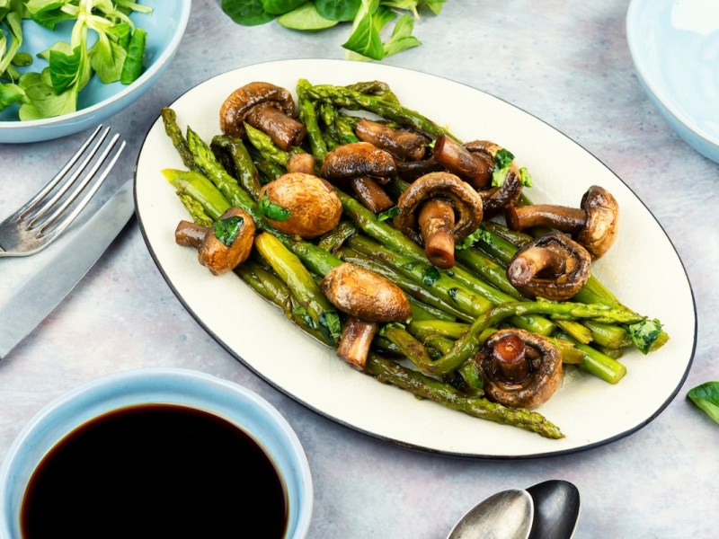 Spargel-Pilz-Pfanne auf einem ovalen Teller, daneben Besteck und eine Schale Feldsalat.