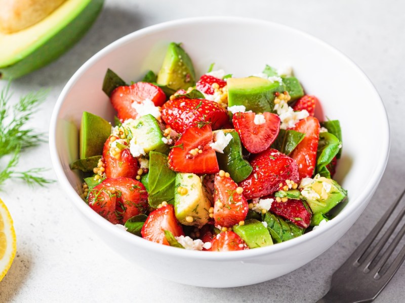 Eine Schüssel Spinatsalat mit Erdbeeren, Avocado und Feta. Daneben eine Gabel, eine halbe Zitrone und eine halbe Avocado. Nahaufnahme.