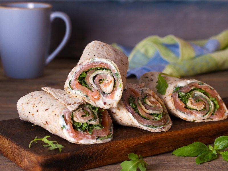4 Wraps mit Lachs und Meerrettich auf einem Holzbrett, dahinter eine Tasse und ein Geschirrtuch.