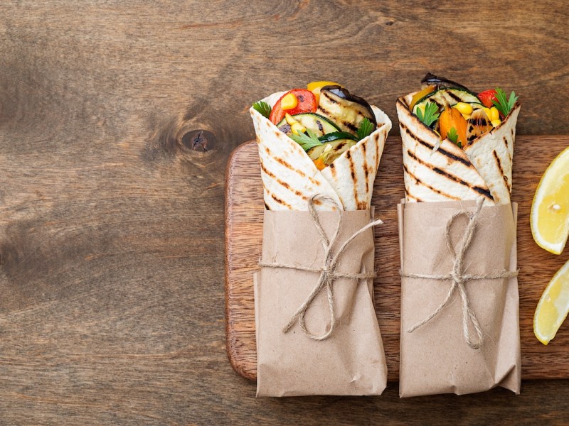 2 Wraps vom Grill gefüllt mit Gemüse und Koriander, eingewickelt in Backpapier, auf einem Brett, daneben zwei Zitronenspalten.