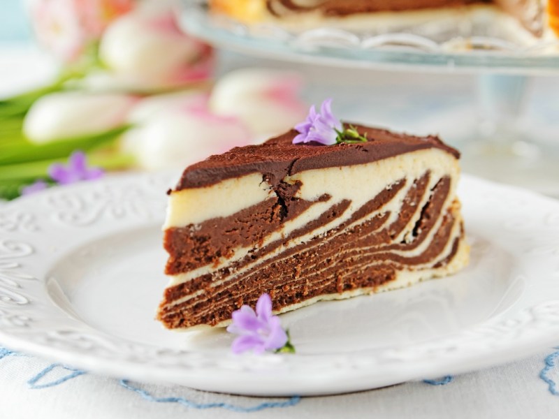 Ein Stück Zebra-Käsekuchen mit hellem und dunklem Teig auf einem Teller, dekoriert mit Blüten.