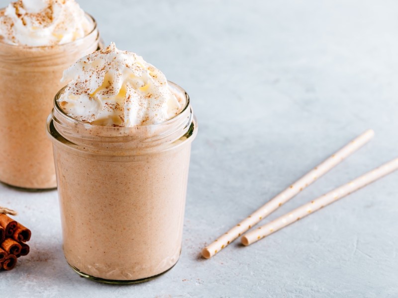 Zwei Gläser Zimtschnecken-Milchshake mit Sahne, daneben Zimtstangen und zwei Strohhalme.