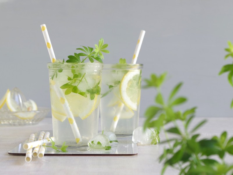 Zwei Gläser mit alkoholfreier Maibowle stehen auf einem weißen Tisch. Vor ihnen liegen ein paar Eiswürfel. Im Vordergrund ist noch frischer Waldmeister.